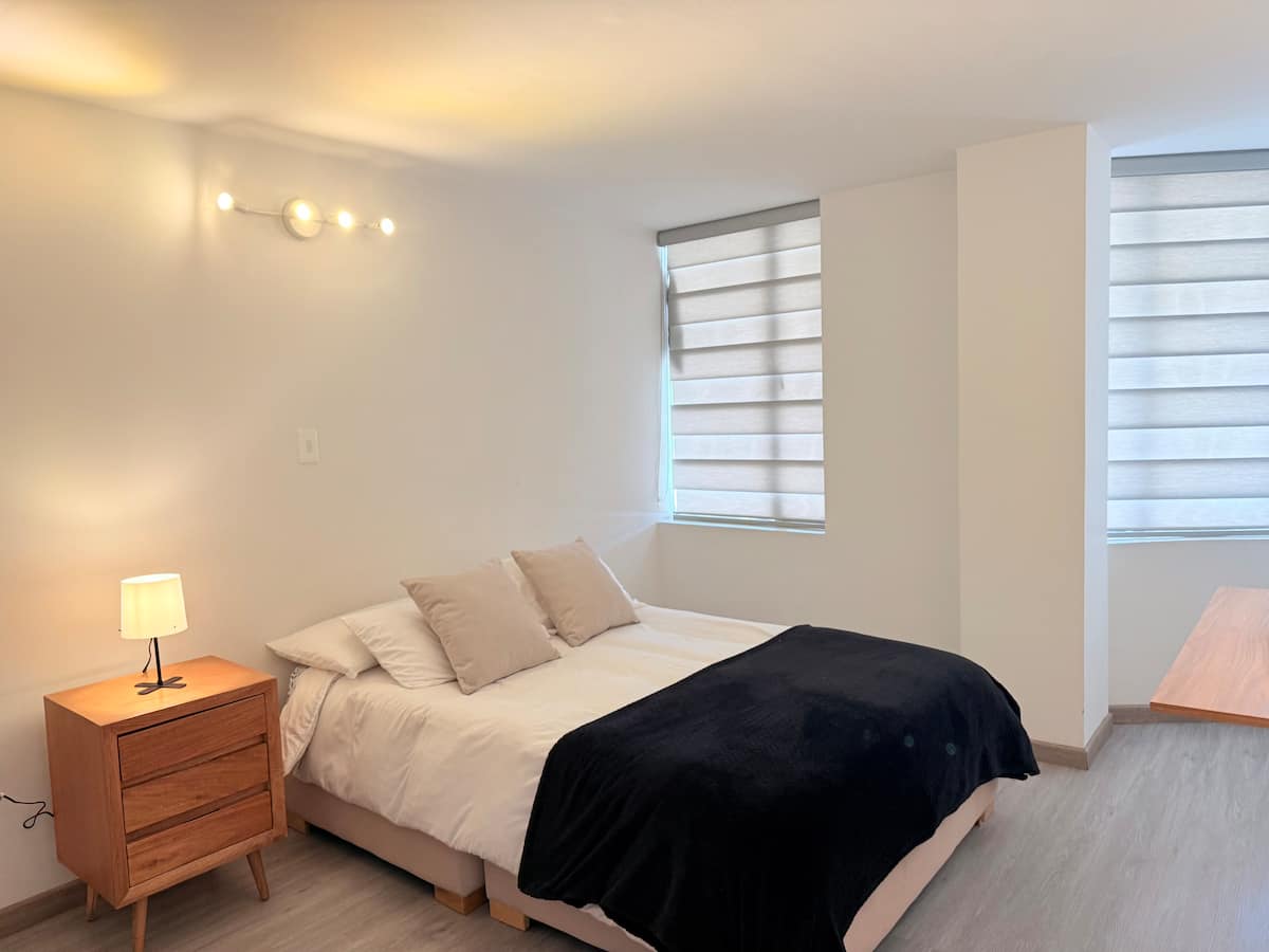 Habitación con cama doble, mesa de noche de madera y dos ventanas con persianas