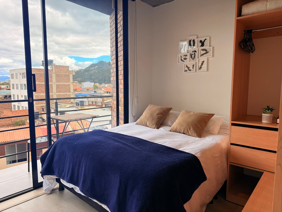 Cama doble con cobija azul frente al ventanal que abre al balcón con vista a Sogamoso