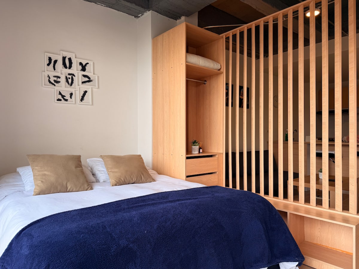 Dormitorio con cama doble, clóset abierto y biombo de listones que separa la cocina
