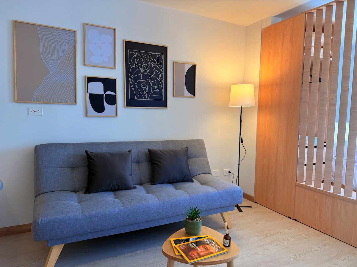 Sofá cama gris bajo una galería de cuadros abstractos, con lámpara de pie encendida