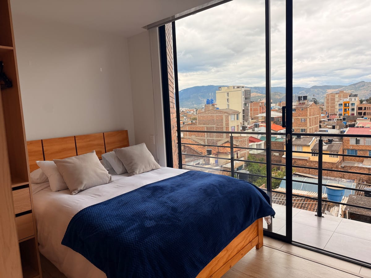 Cama doble con cobija azul frente al ventanal corredizo con vista a la ciudad