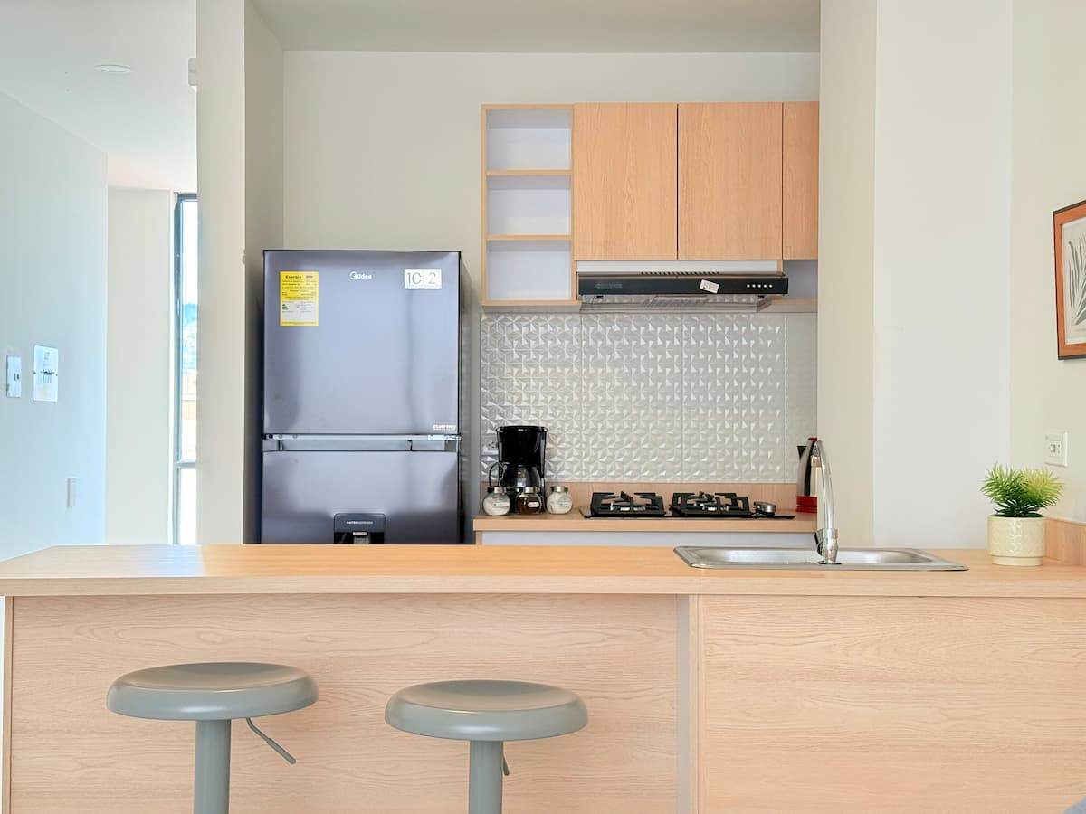 Cocina abierta con barra de madera, dos taburetes, estufa a gas y nevera gris
