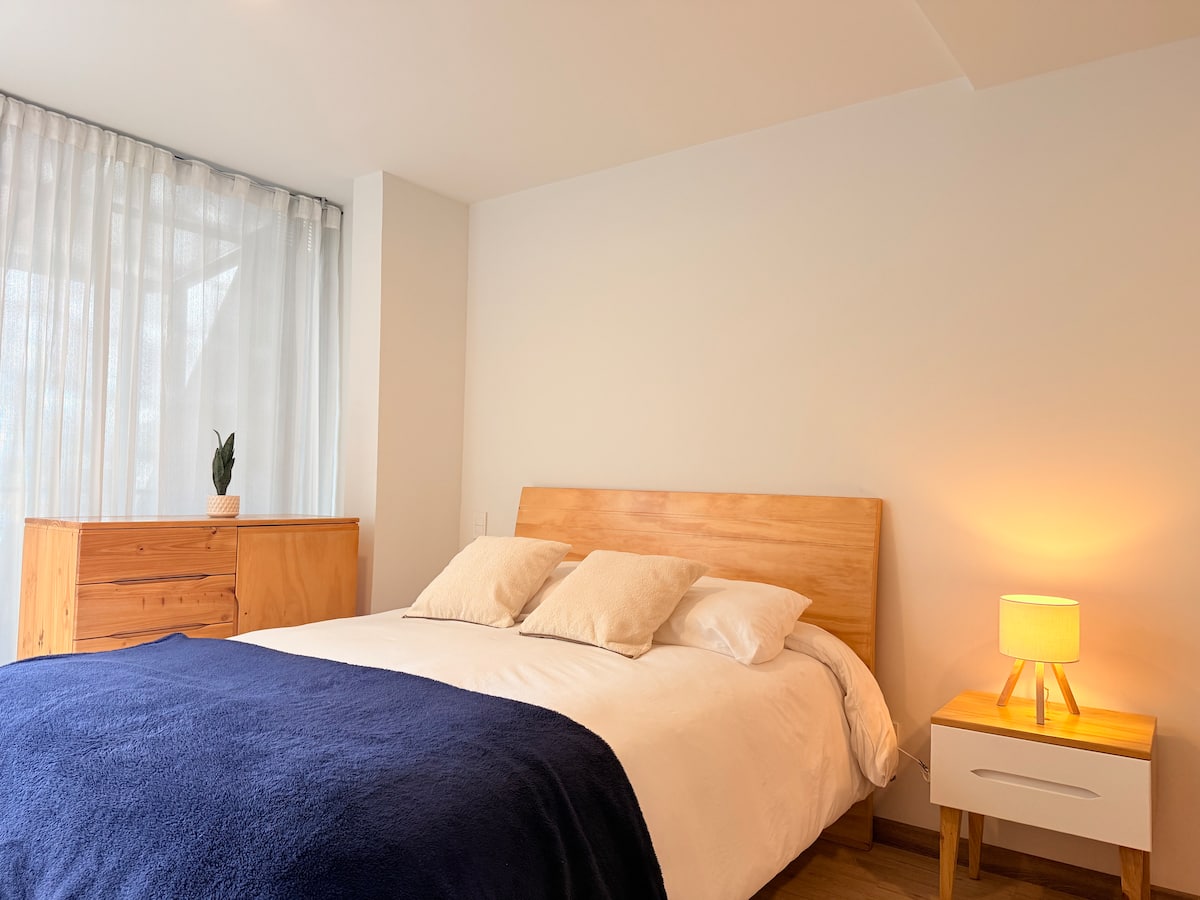 Cama doble con cobija azul, cortinas blancas translúcidas y lámpara de noche encendida