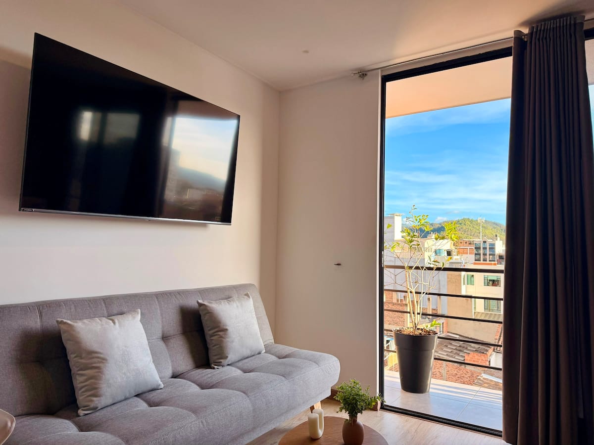 Televisor en la pared sobre el sofá gris, con la puerta abierta al balcón soleado