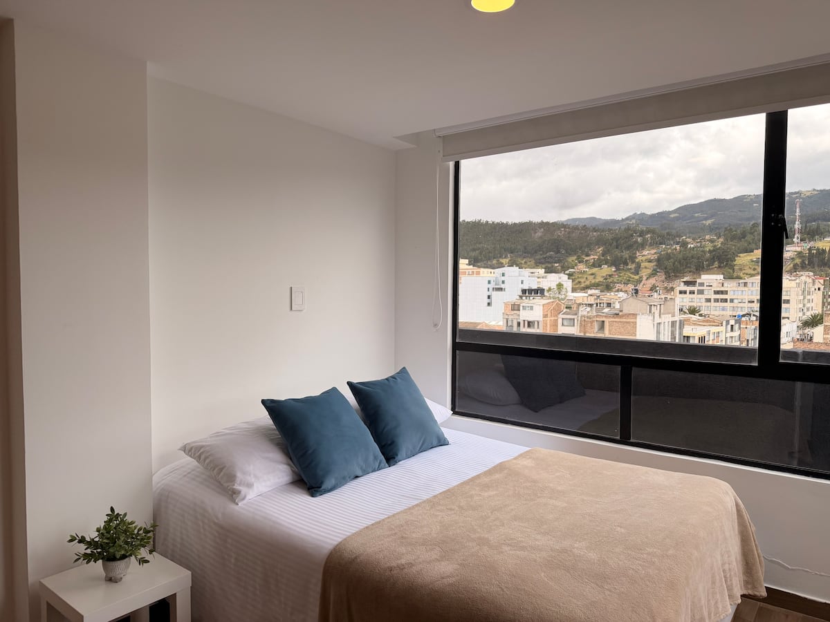 Habitación con cama doble, cojines azules y ventanal con vista a la ciudad y los cerros